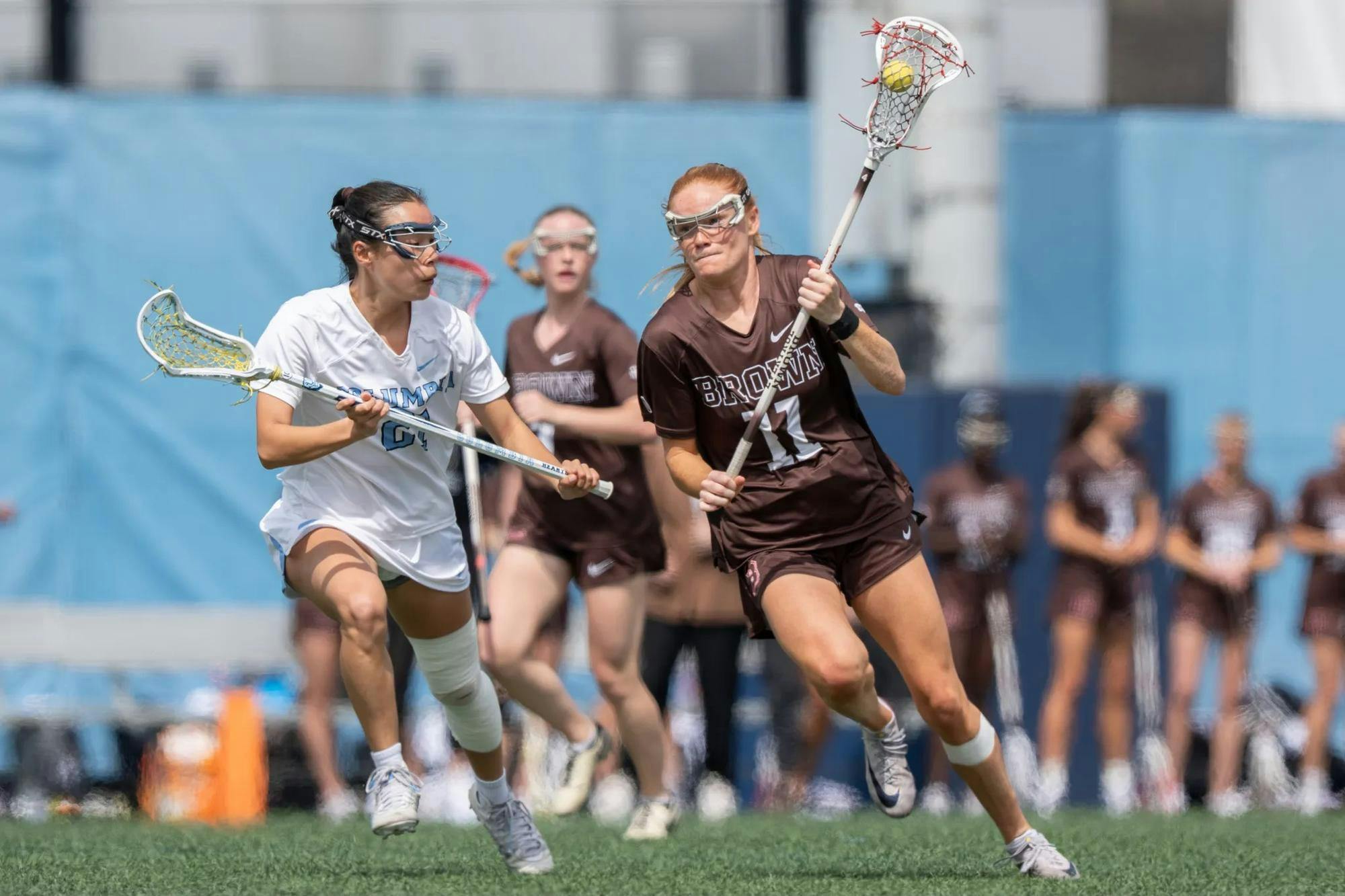 Photo of a Brown lacrosse player running while holding up a ball with a netted stick, accompanied by a competing Columbia player to her left. The blurred background features another Brown player on the field, a resting group of athletes and a blue backdrop.