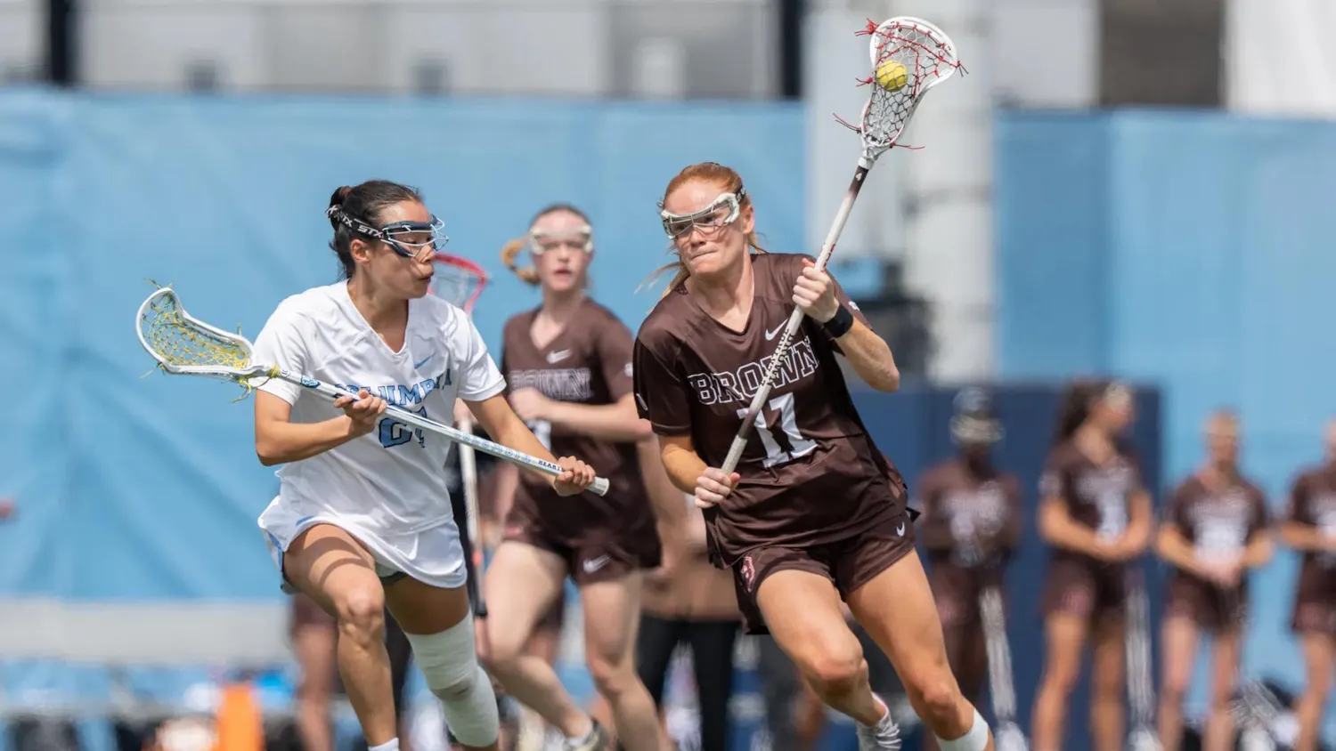 Photo of a Brown lacrosse player running while holding up a ball with a netted stick, accompanied by a competing Columbia player to her left. The blurred background features another Brown player on the field, a resting group of athletes and a blue backdrop.