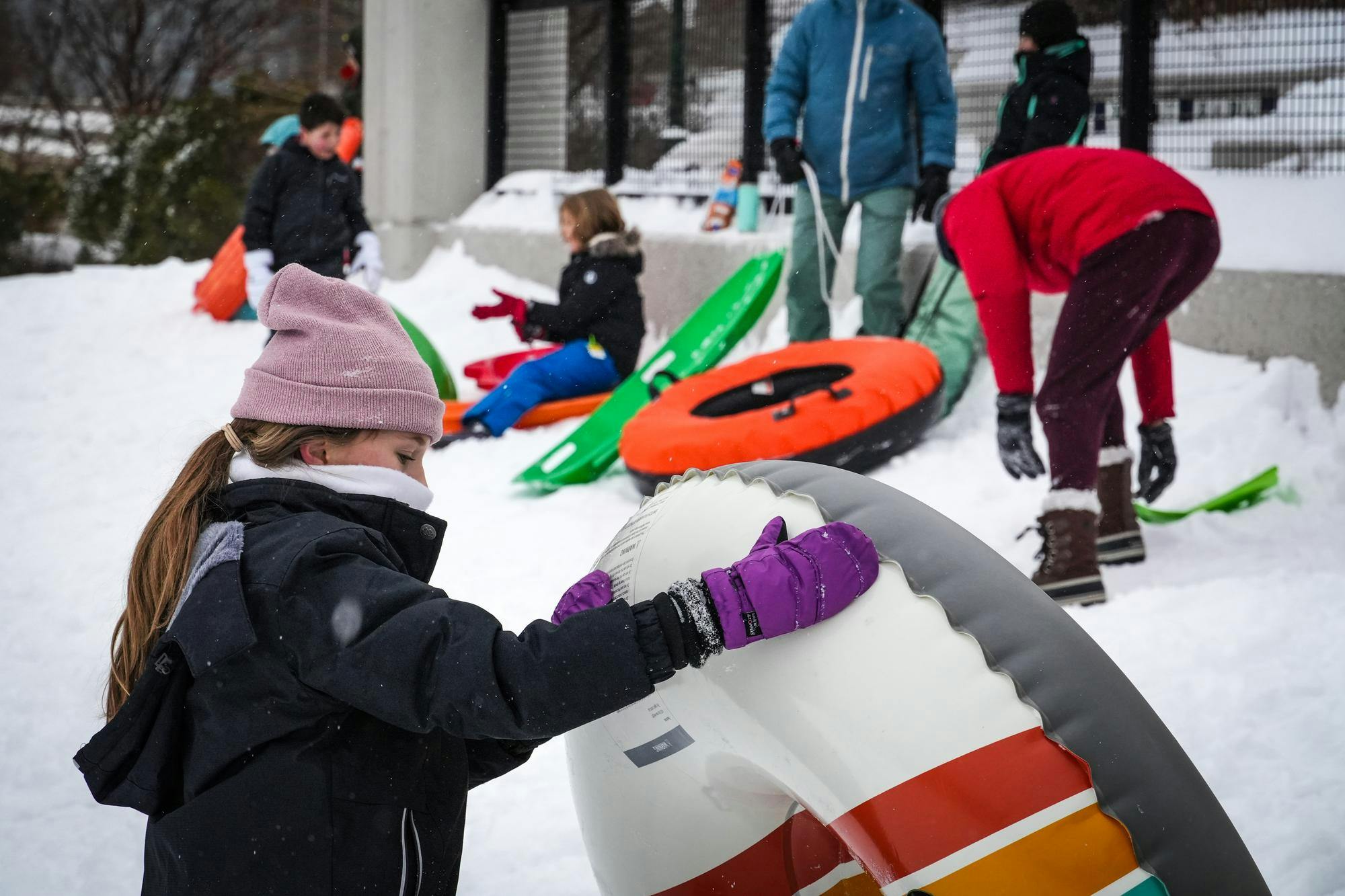 A little girl with a pink hat and black jacket holds a snow tube.