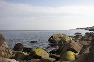 A photo of the Rhode Island coast shows rocks lining a beach that extends along the horizon.