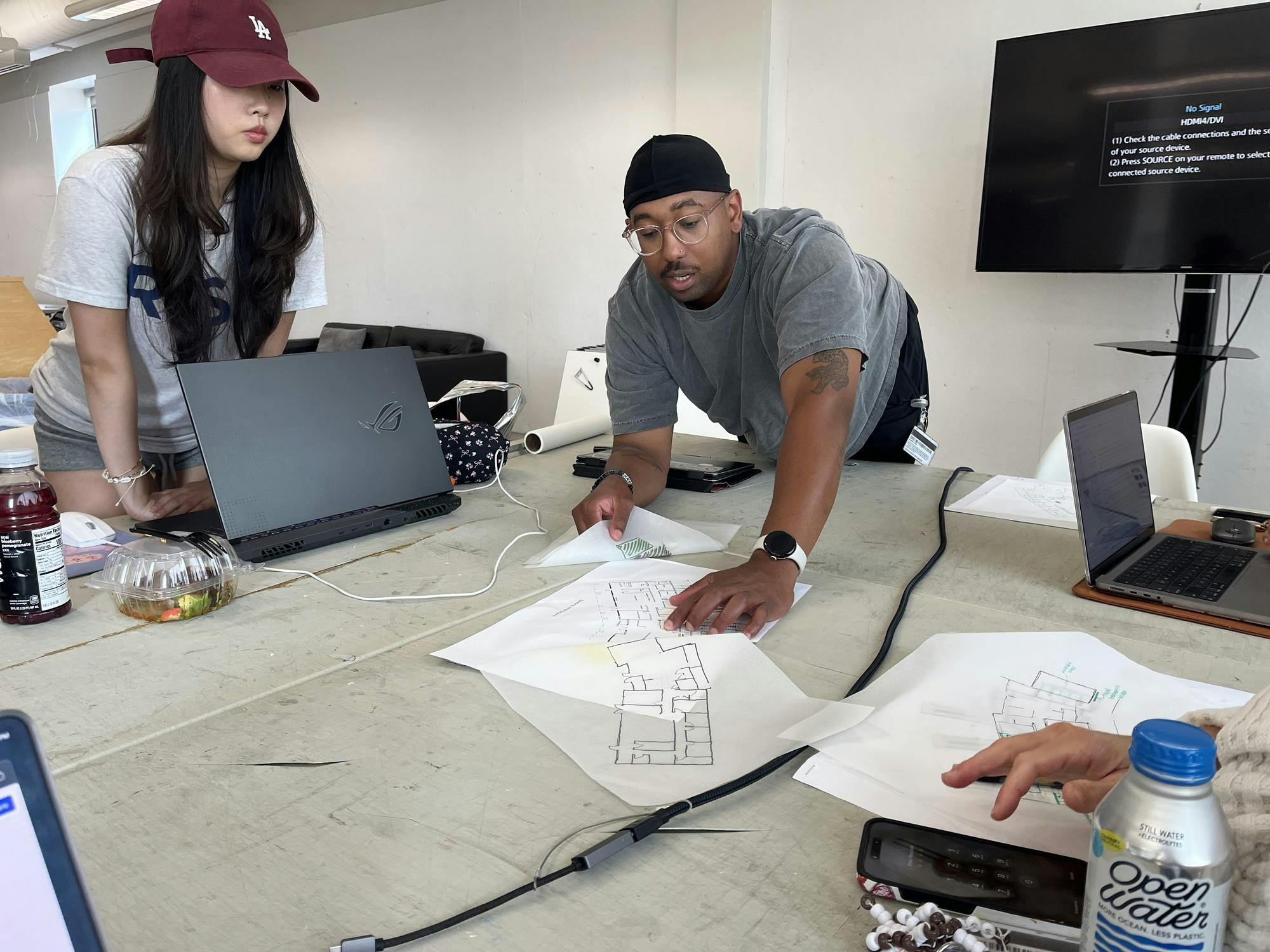 Photo of someone leaning over a table to point at a floor-plan drawing on tracing paper as someone to his right looks over their laptop to look as well.