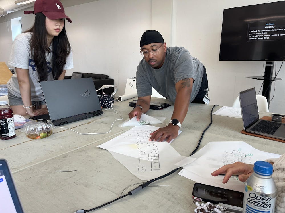 Photo of someone leaning over a table to point at a floor-plan drawing on tracing paper as someone to his right looks over their laptop to look as well.