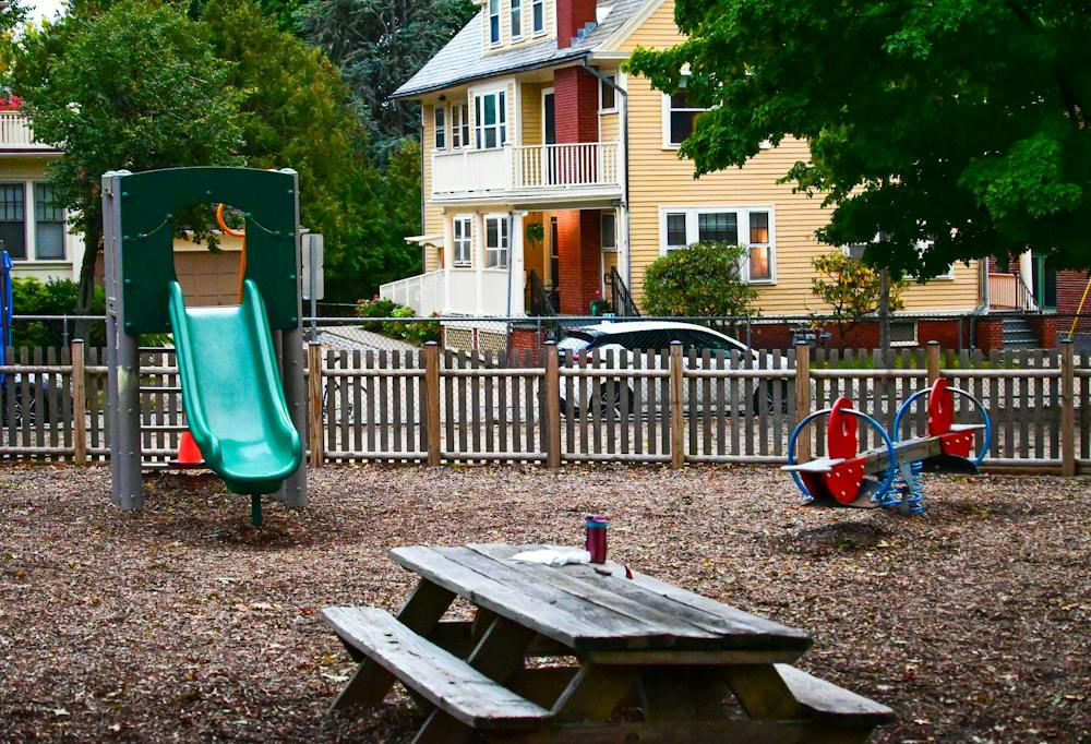A photo of a playground with a green slide and a red and blue see-saw in addition to a wooden table with benches surrounded by a wooden fence.