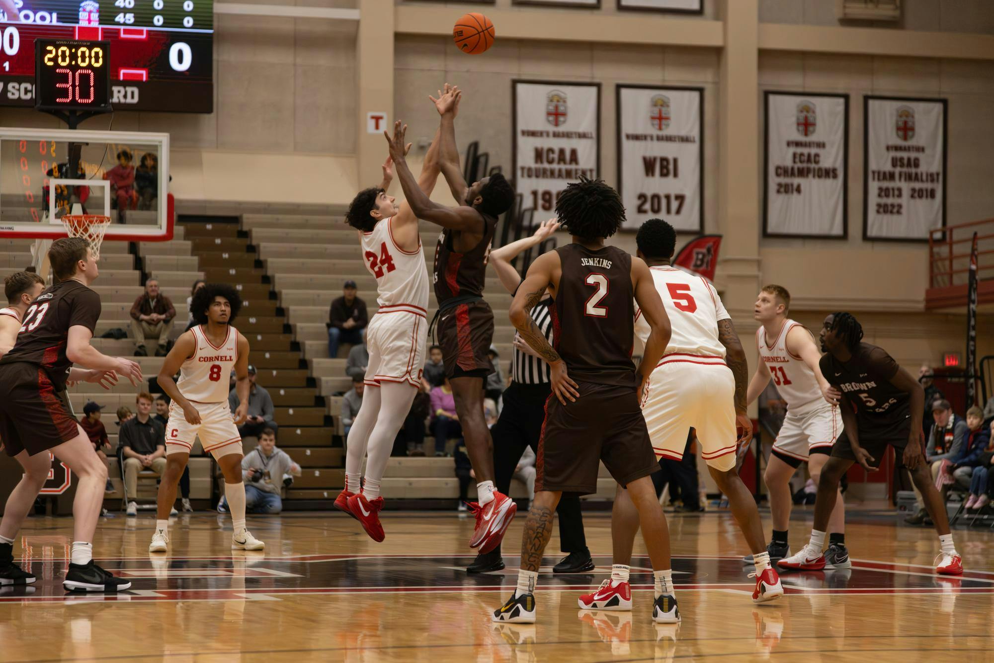 The opening tip-off of a basketball game. Two players, one in white and red and the other in brown and red, are mid-jump, both reaching for the ball. 