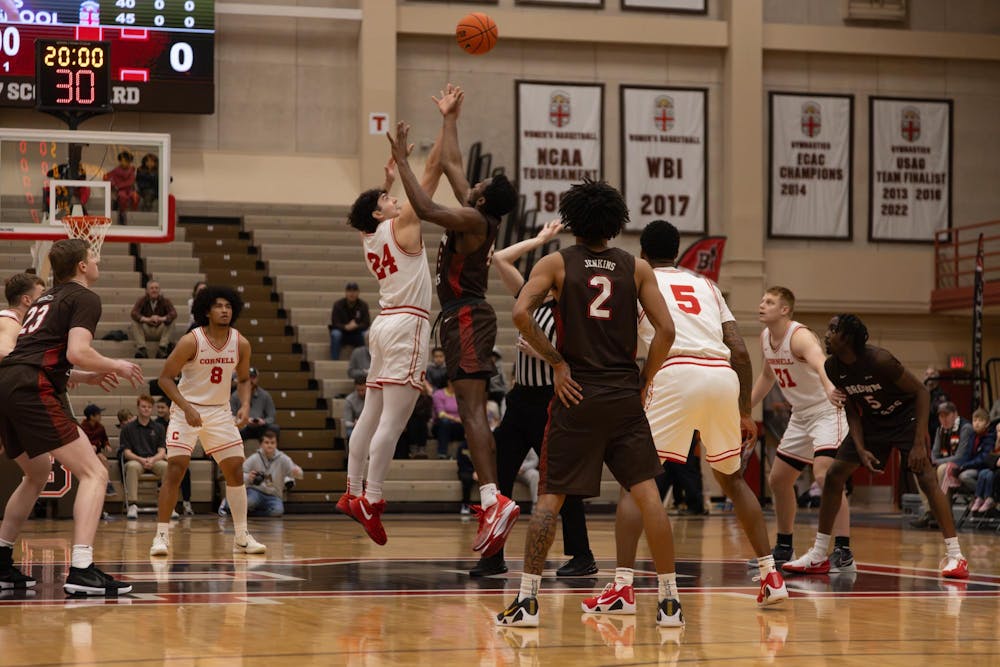 The opening tip-off of a basketball game. Two players, one in white and red and the other in brown and red, are mid-jump, both reaching for the ball. 