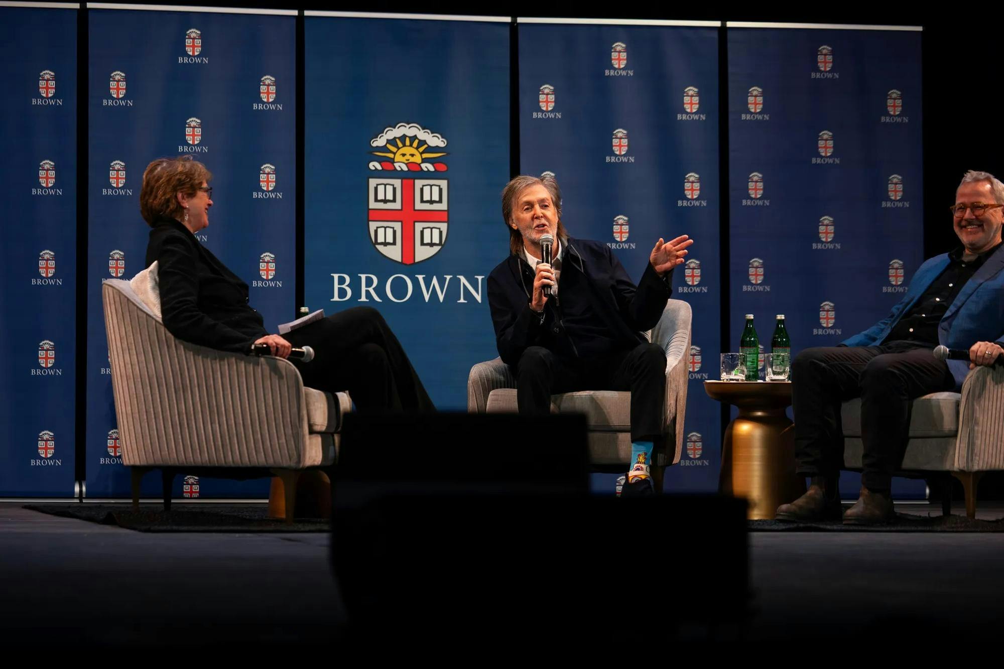 Photo of the guest panel featuring Christina Paxson on the left, Paul McCartney in the middle speaking into the microphone and Morgan Neville on the right, all sat in gray chairs against a blue backdrop with the Brown University seals. 