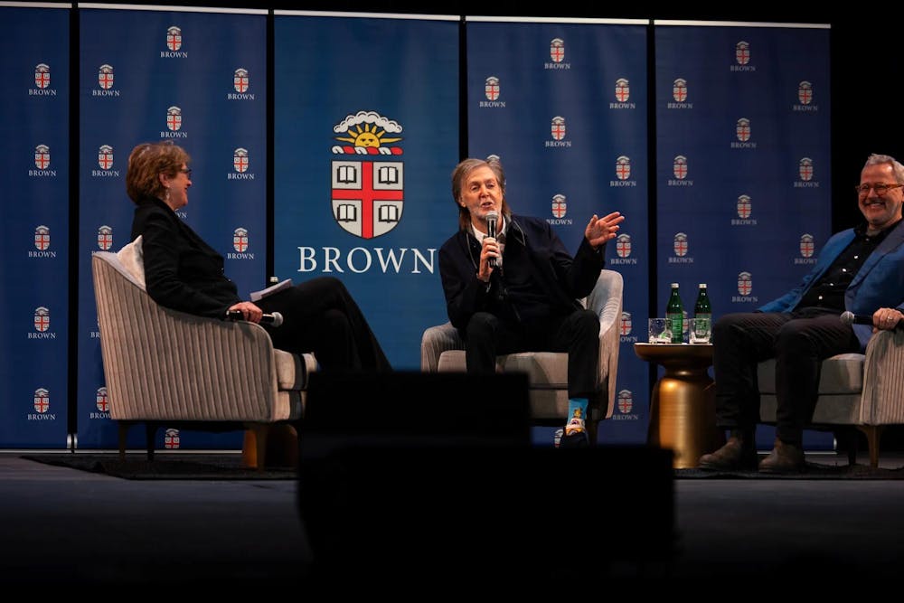 Photo of the guest panel featuring Christina Paxson on the left, Paul McCartney in the middle speaking into the microphone and Morgan Neville on the right, all sat in gray chairs against a blue backdrop with the Brown University seals. 