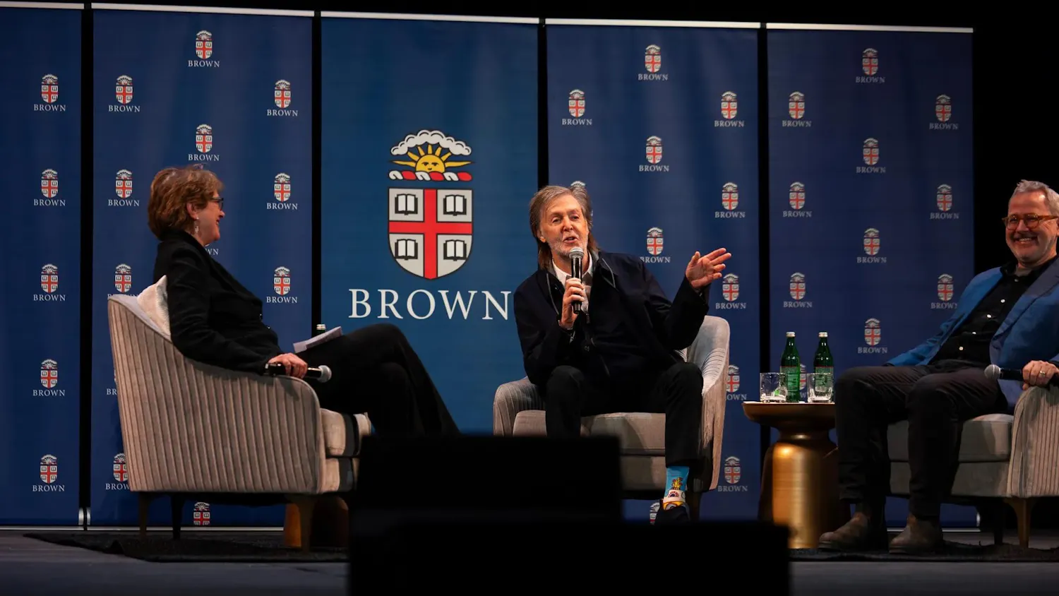 Photo of the guest panel featuring Christina Paxson on the left, Paul McCartney in the middle speaking into the microphone and Morgan Neville on the right, all sat in gray chairs against a blue backdrop with the Brown University seals.