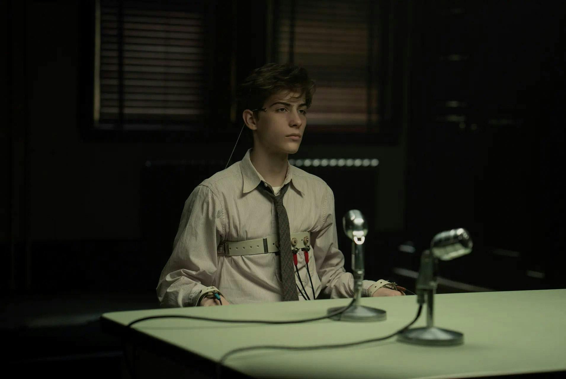 Frame from the movie, “Unabomber”, showing a young boy wearing a shirt and tie, with straps along his chest and wrists, sitting at a table with a microphone positioned in front of him atop a table.
