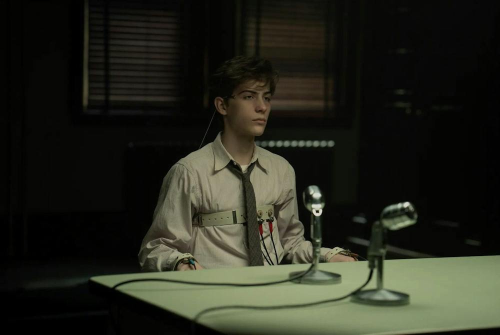 Frame from the movie, “Unabomber”, showing a young boy wearing a shirt and tie, with straps along his chest and wrists, sitting at a table with a microphone positioned in front of him atop a table.