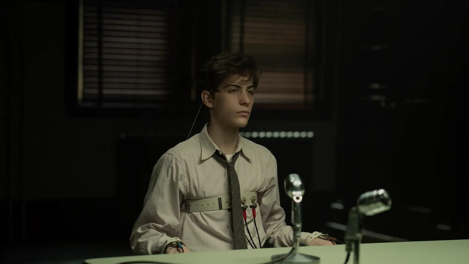 Frame from the movie, “Unabomber”, showing a young boy wearing a shirt and tie, with straps along his chest and wrists, sitting at a table with a microphone positioned in front of him atop a table.