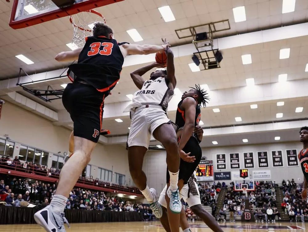 A Brown basketball player mid-air, about to score a point, while two Princeton players attempt to block him. 

