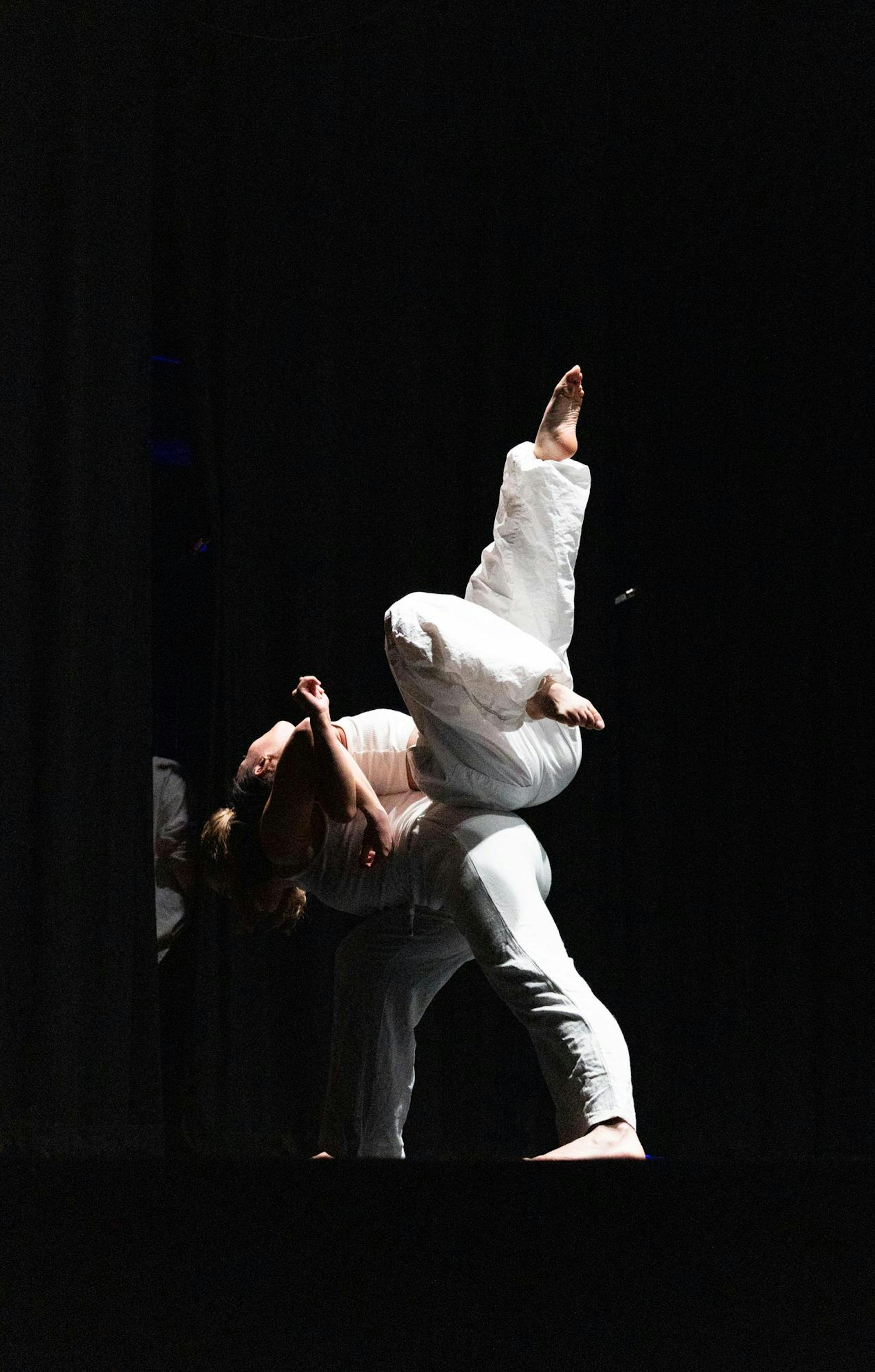 Two dancers in all white back to back with one balancing the other. 