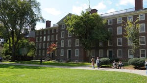 A photo of Brown University's University Hall, taken from the Main Green. Two people sit on the bench in front of the building. Three people walk on the sidewalk in front of the building.