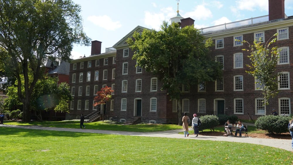 A photo of Brown University's University Hall, taken from the Main Green. Two people sit on the bench in front of the building. Three people walk on the sidewalk in front of the building.