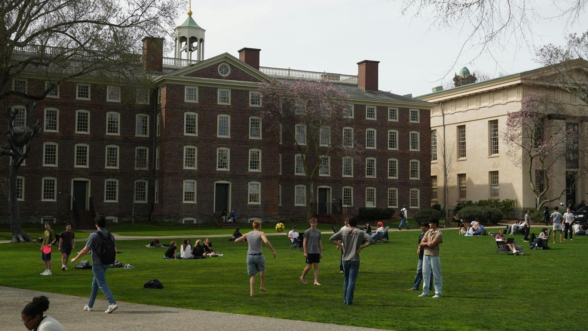 A photo of the Main Green with University Hall in the background. A group of students are playing with a soccer ball while others are sitting on chairs and blankets. 