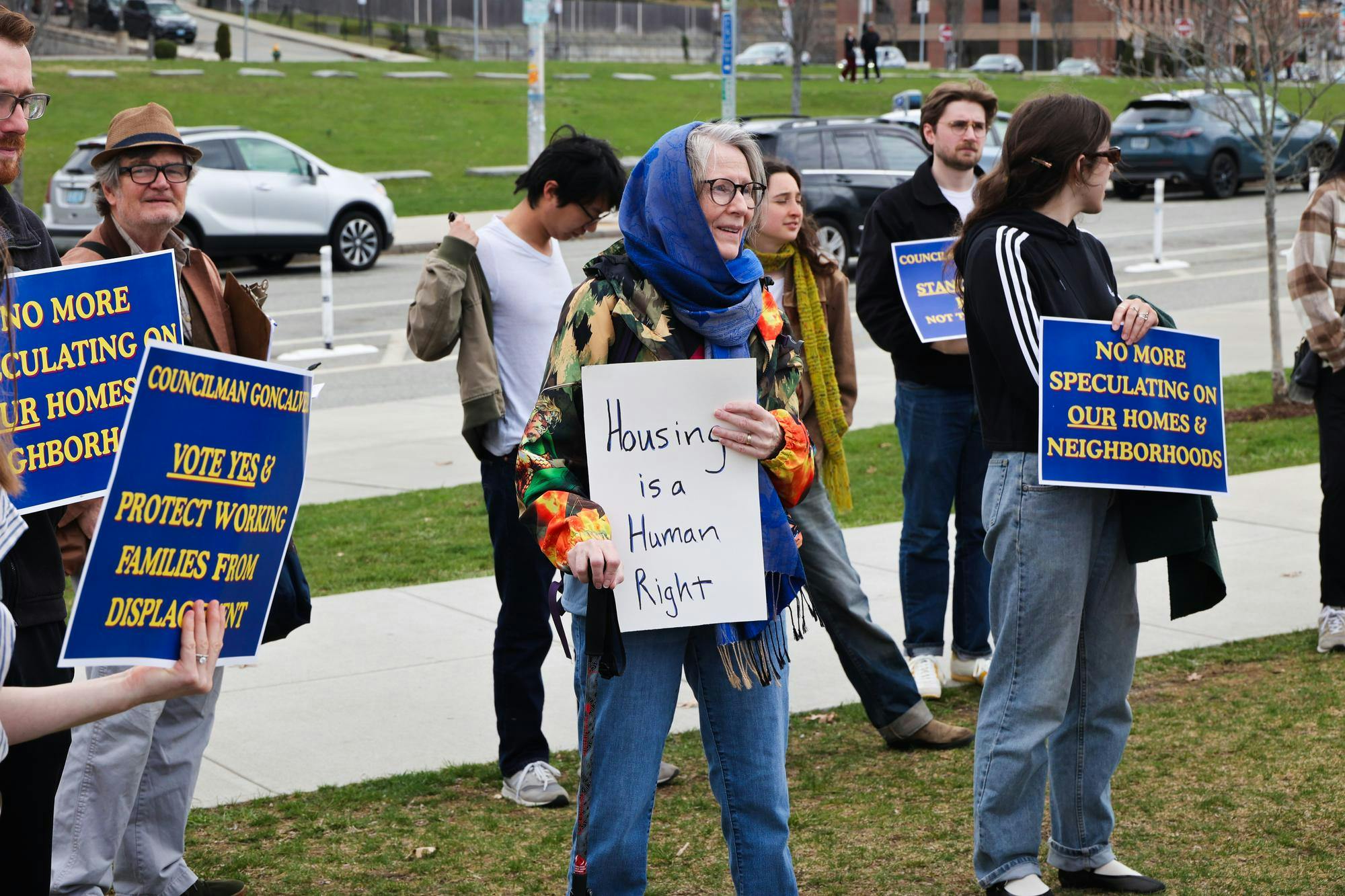 A photo of rallyers standing in the grass along a main road holding signs calling for rent stabilization. 
