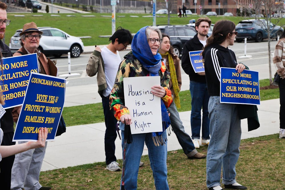 A photo of rallyers standing in the grass along a main road holding signs calling for rent stabilization. 
