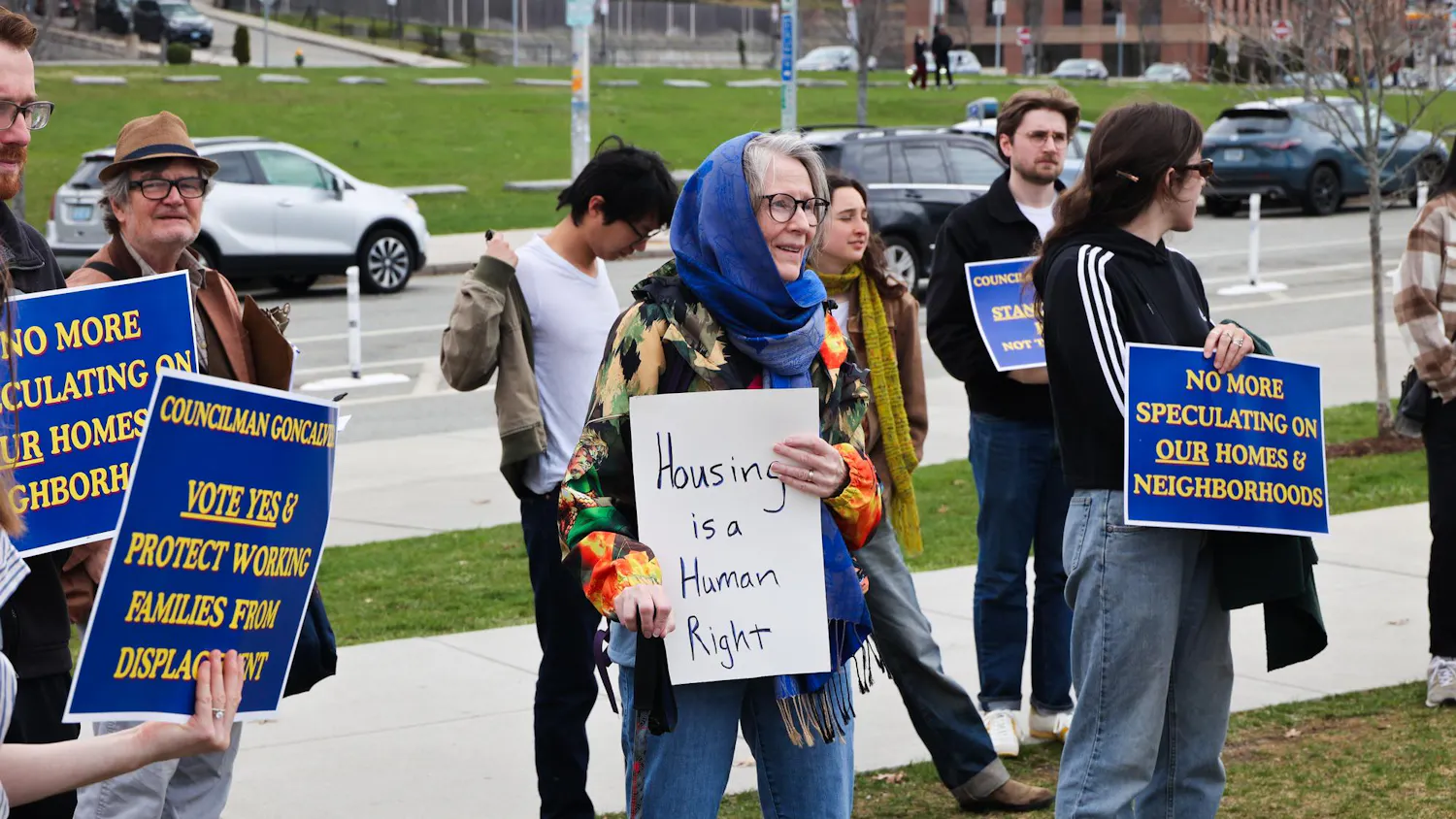 A photo of rallyers standing in the grass along a main road holding signs calling for rent stabilization.