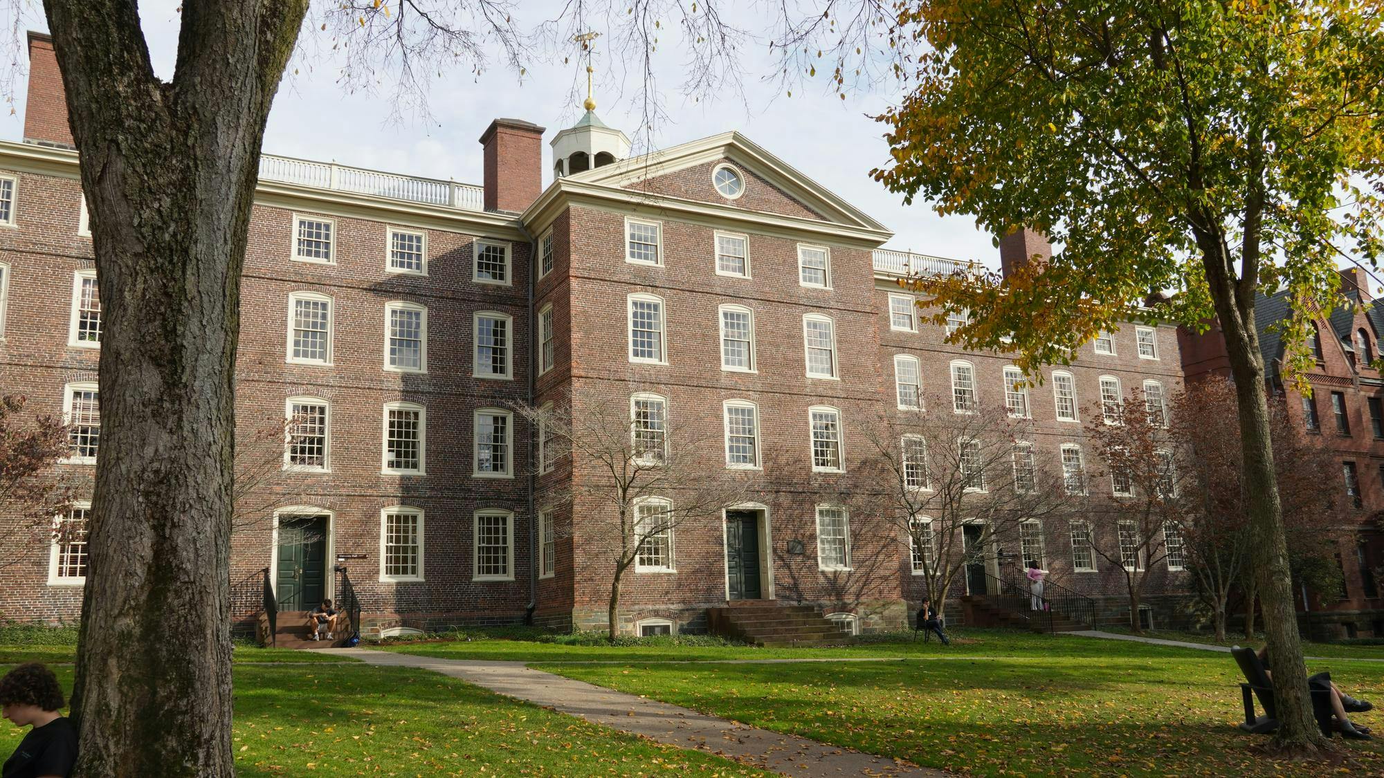 Photo of the University Hall with surrounding trees, pathways around the front yard and multiple students scattered around. 