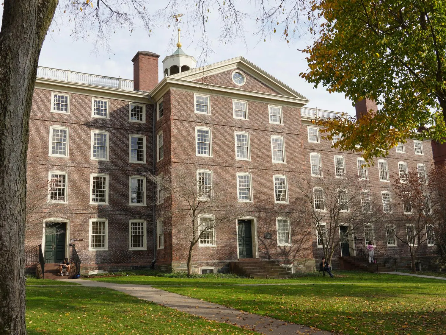 Photo of the University Hall with surrounding trees, pathways around the front yard and multiple students scattered around.