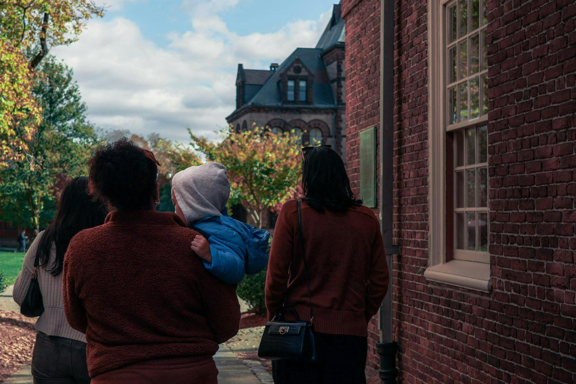Photo of three adult individuals walking to the left of a brick building onto the Main Green, one of whom is carrying a baby in a blue coat with a grey hood.
