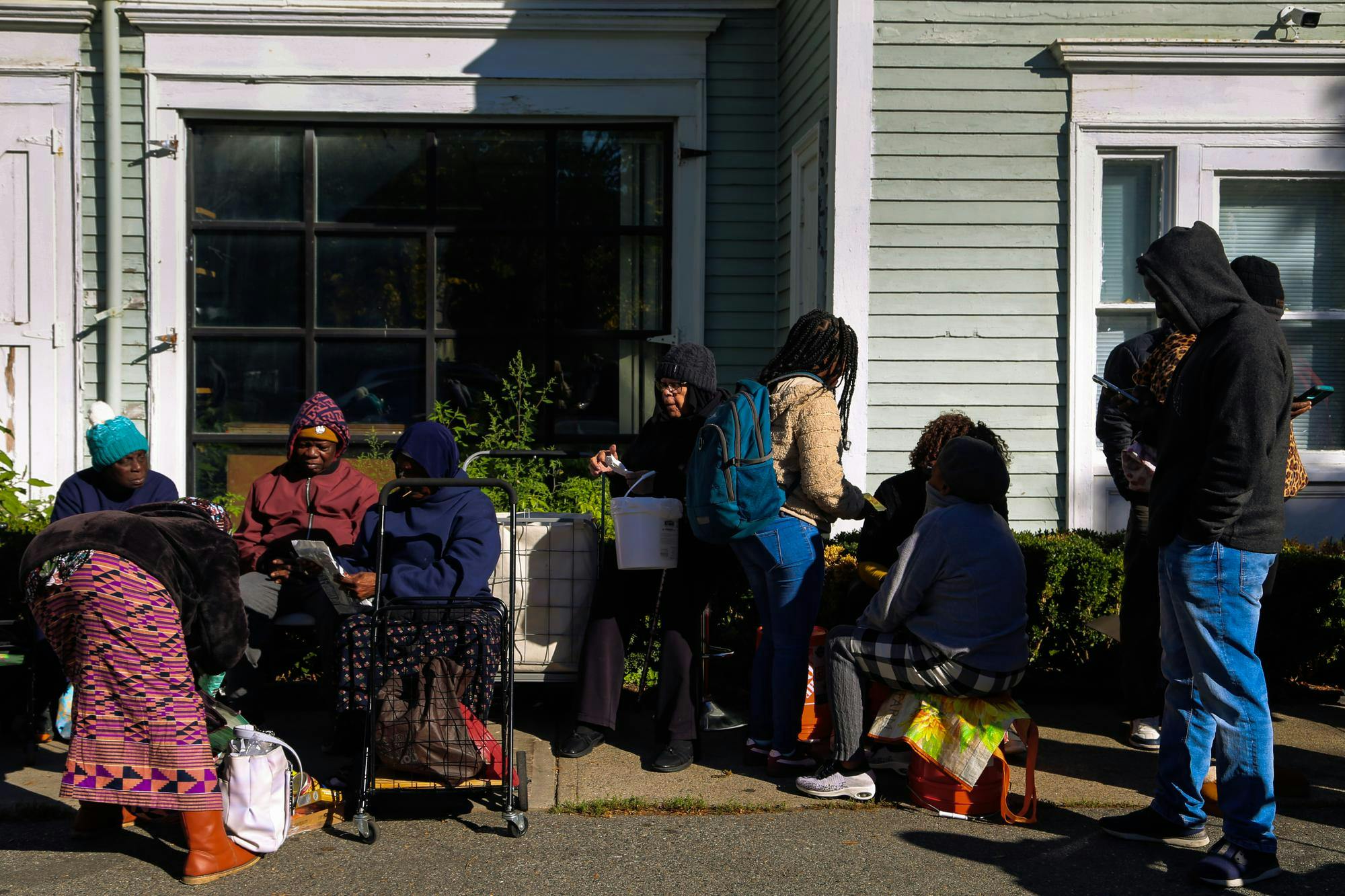 Photo of people in a food distribution line wearing hoodies, hats and sweaters while carrying carts and backpacks on a sunny day in front of Women's Refugee Care.