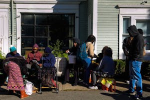 Photo of people in a food distribution line wearing hoodies, hats and sweaters while carrying carts and backpacks on a sunny day in front of Women's Refugee Care.