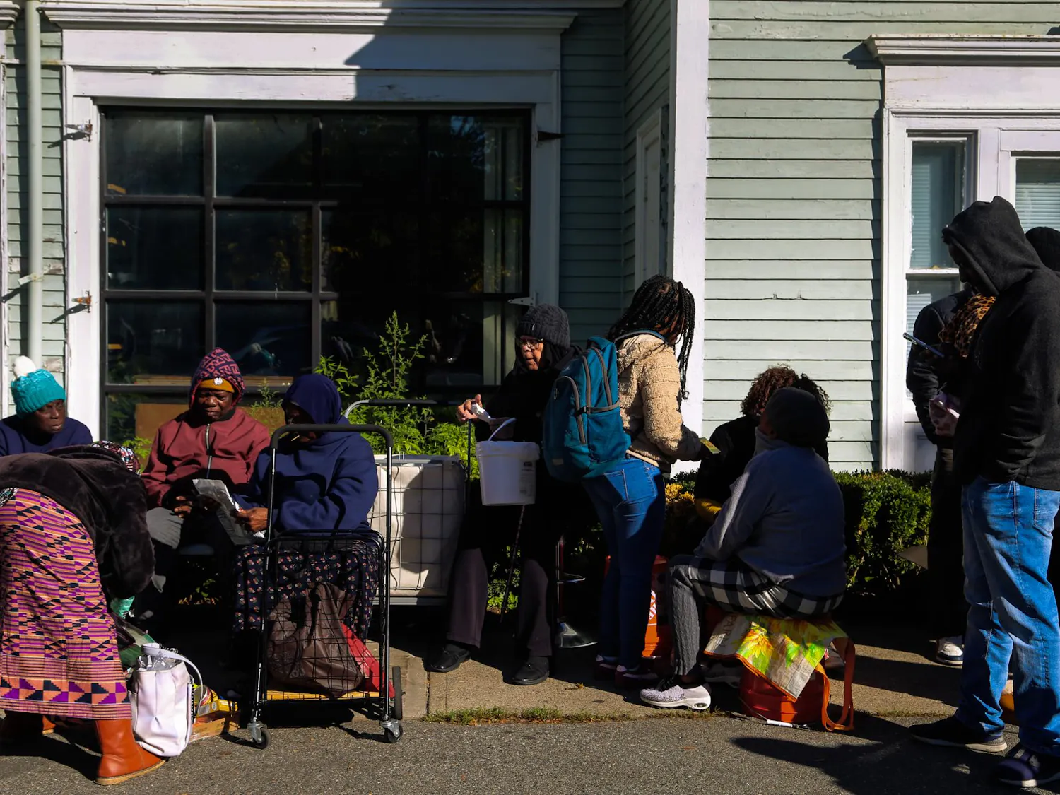 Photo of people in a food distribution line wearing hoodies, hats and sweaters while carrying carts and backpacks on a sunny day in front of Women's Refugee Care.