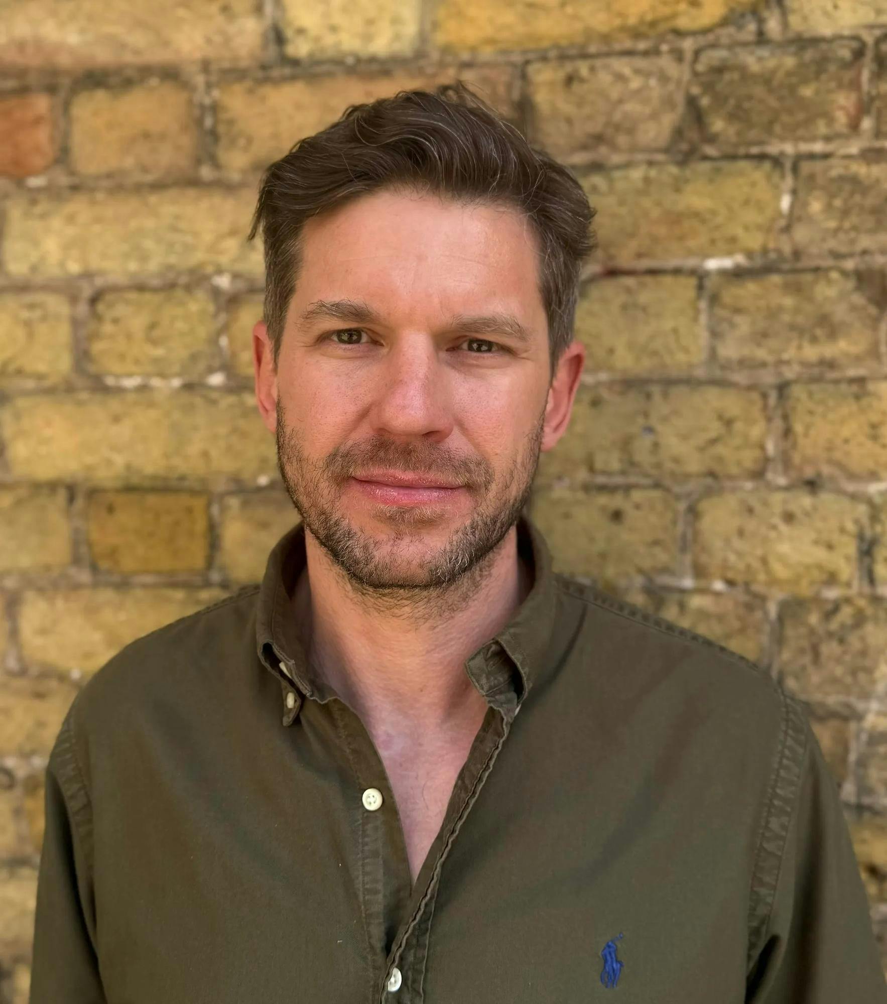 Photo of a man with a beard and mustache wearing an army green button down shirt with the top three buttons unbuttoned standing in front of a yellow brick wall. 