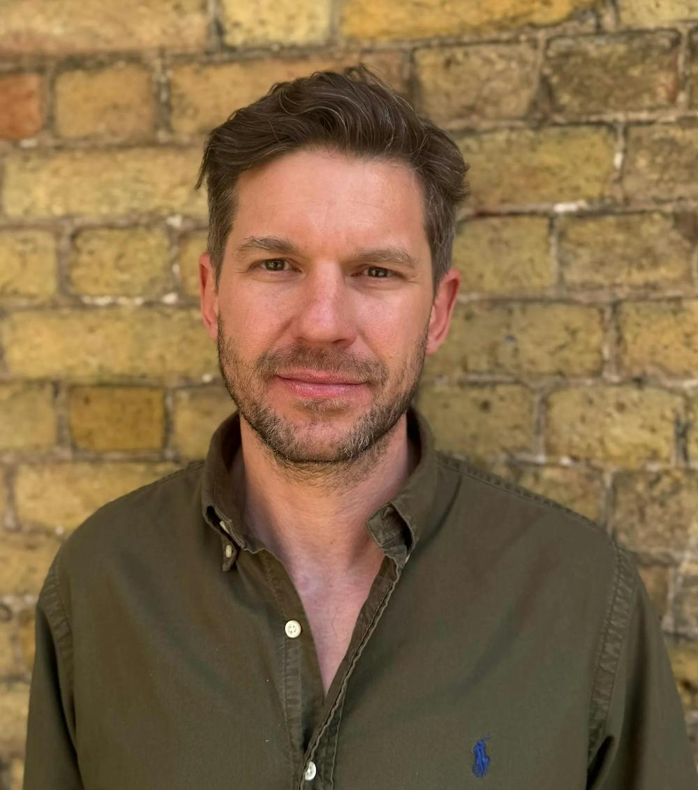 Photo of a man with a beard and mustache wearing an army green button down shirt with the top three buttons unbuttoned standing in front of a yellow brick wall. 