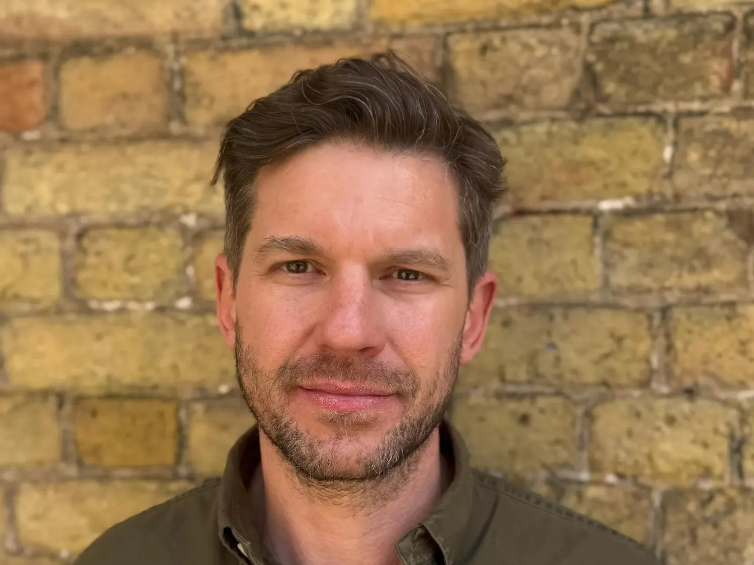 Photo of a man with a beard and mustache wearing an army green button down shirt with the top three buttons unbuttoned standing in front of a yellow brick wall.