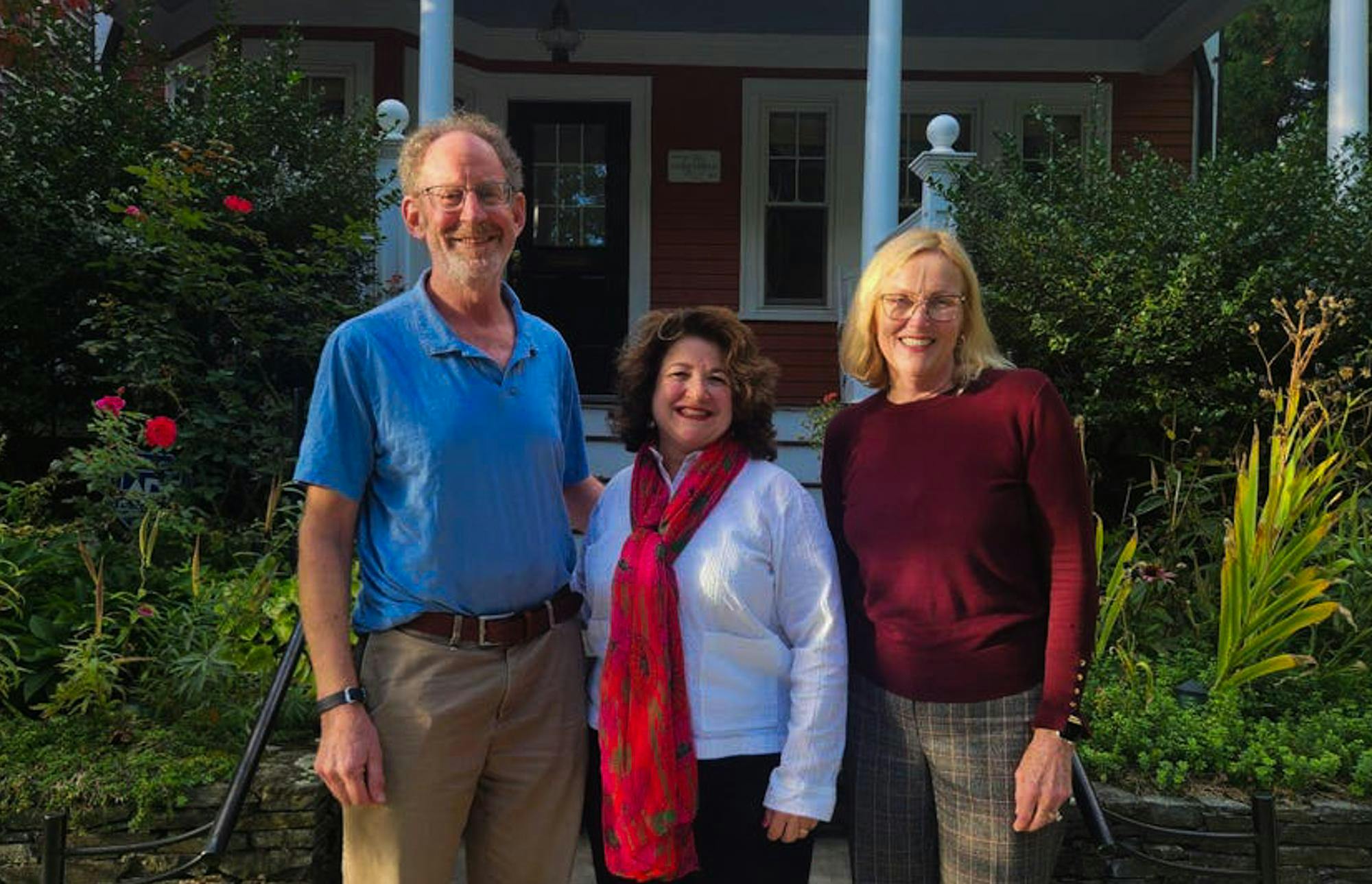 Left: a smiling man wearing a light blue collared shirt, khaki pants, and a brown belt. Middle: a smiling woman wearing a white long sleeved top with a long red scarf. Right: a smiling woman wearing a dark red long-sleeved top with grey patterned pants.