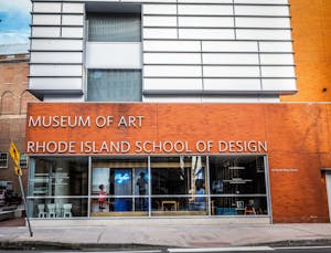 A photo of RISD's Museum of Art, a large orange brick building with large glass windows featuring artwork covering half of the front of the museum. The Photo is taken from across the street, facing the front of the museum. 