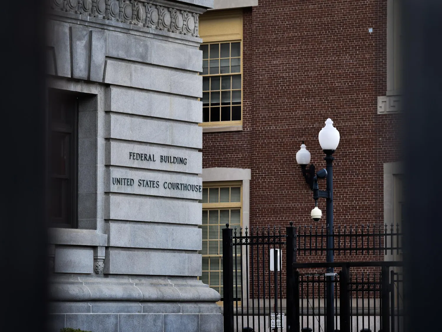 Photo of the U.S. District Court of Rhode Island viewed through two black fence bars. Signage on the courthouse wall reads "Federal Building United States Courthouse."