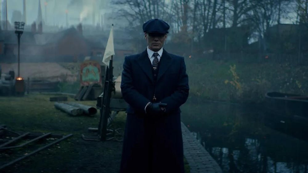 Photo of Tommy Shelby (Cillian Murphy) from Peaky Blinders in a long navy trench coat and cap.

