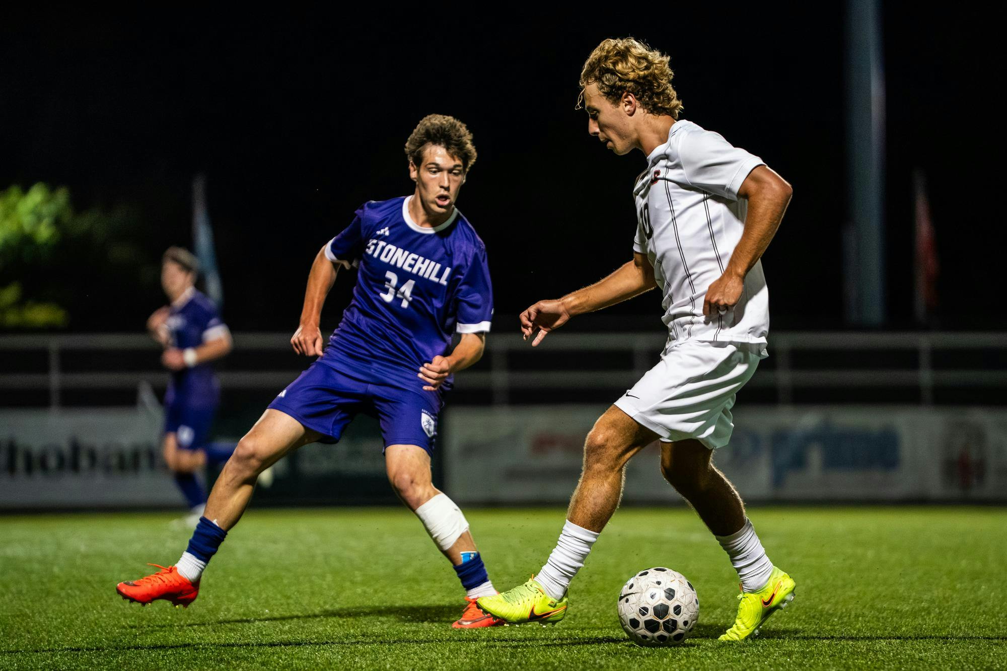 A photo of a Brown University soccer player in a white jersey defending a ball from a Stonehill soccer player wearing a blue jersey on the field at night. 