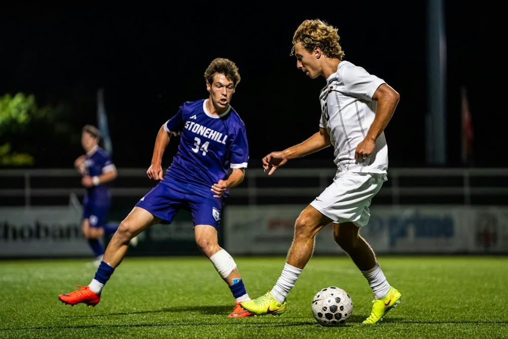 A photo of a Brown University soccer player in a white jersey defending a ball from a Stonehill soccer player wearing a blue jersey on the field at night. 