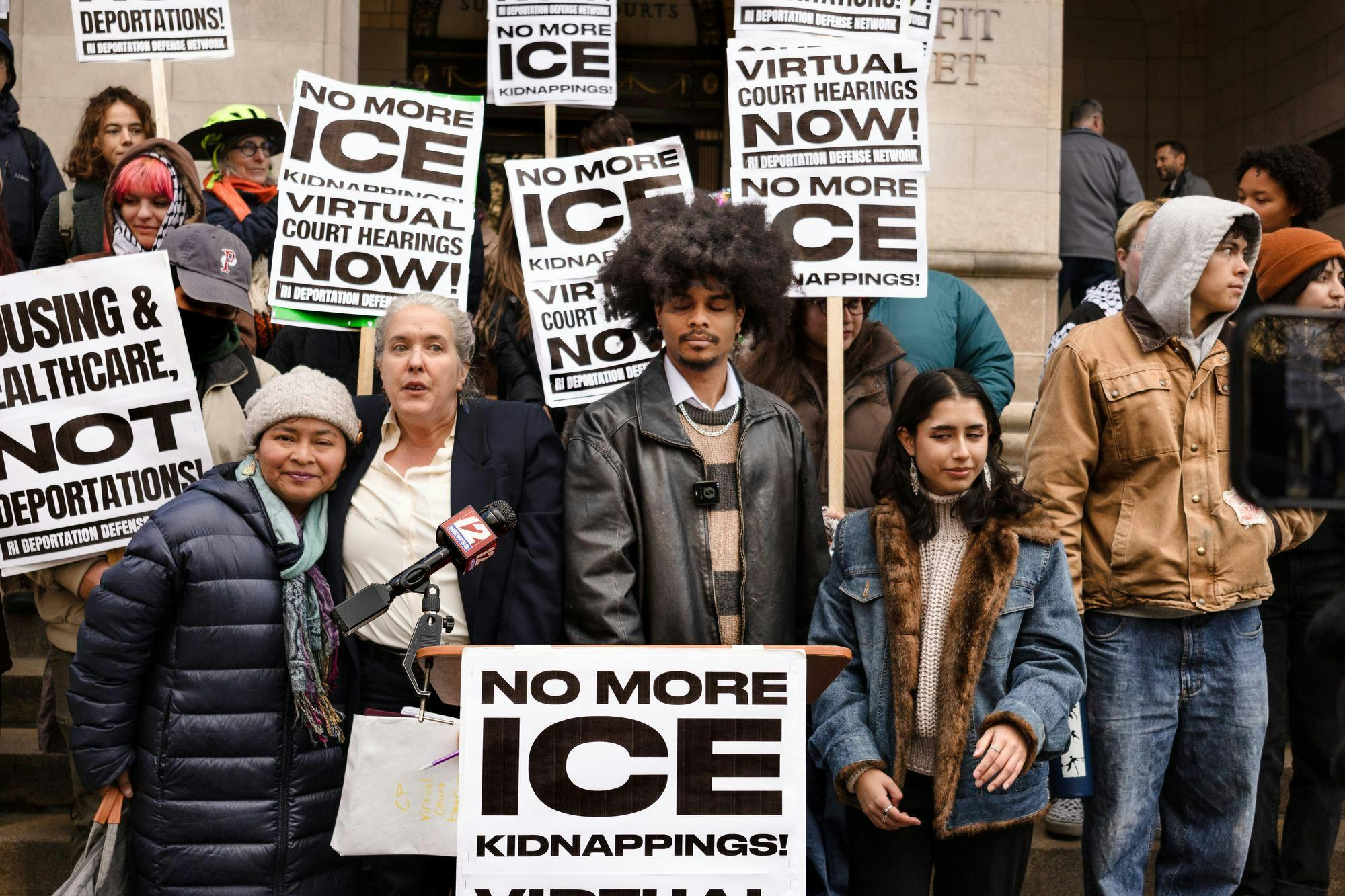 A group of people in front of a podium outside the courthouse. Signs reading "No more ice."