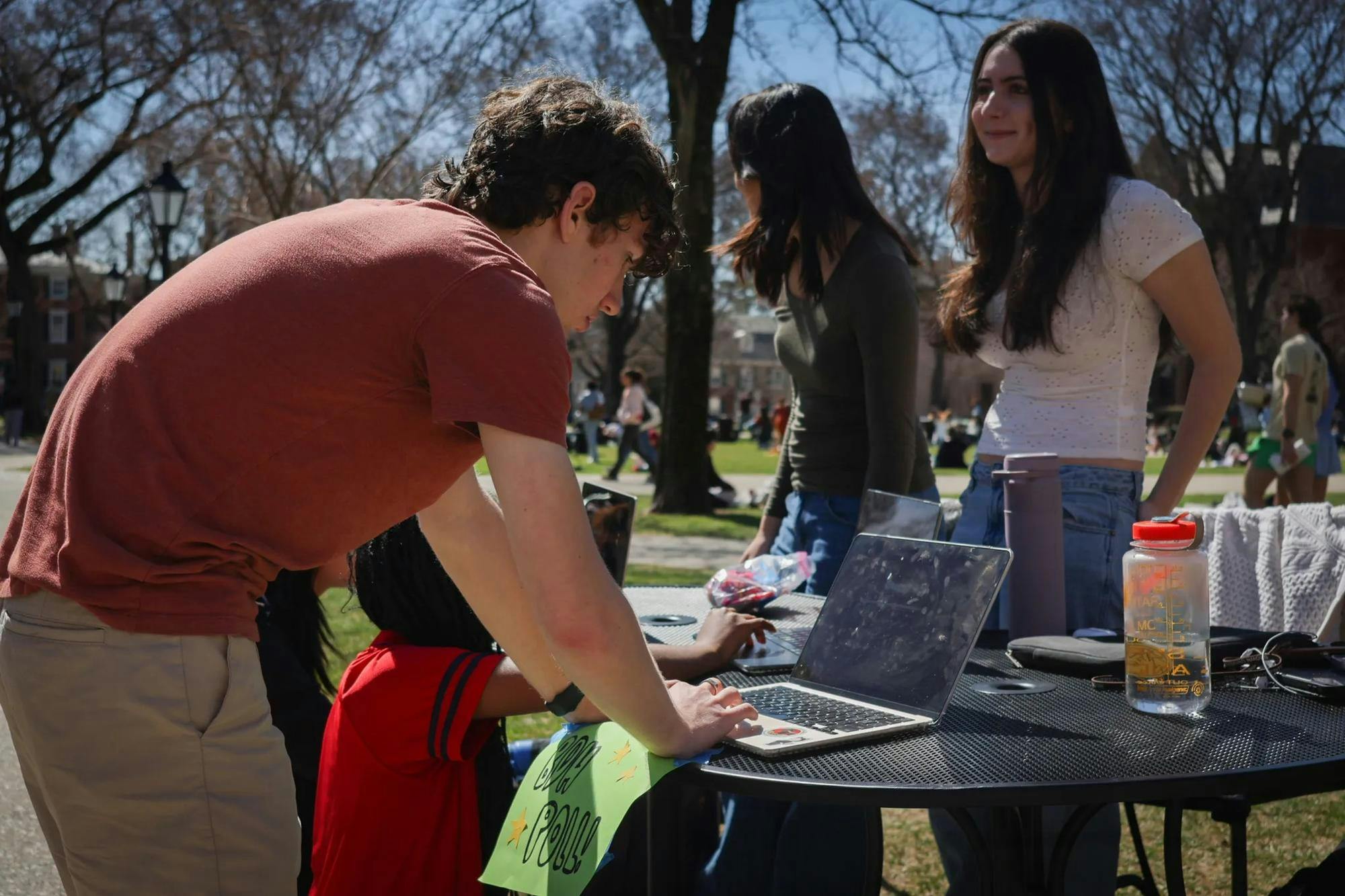 A male undergraduate with curly brown hair is filling out The Herald's spring poll at a polling station on the Main Green. The sun is shining, and two Herald staffers, one wearing a white blouse, the other a dark green shirt, are standing behind the polling station.