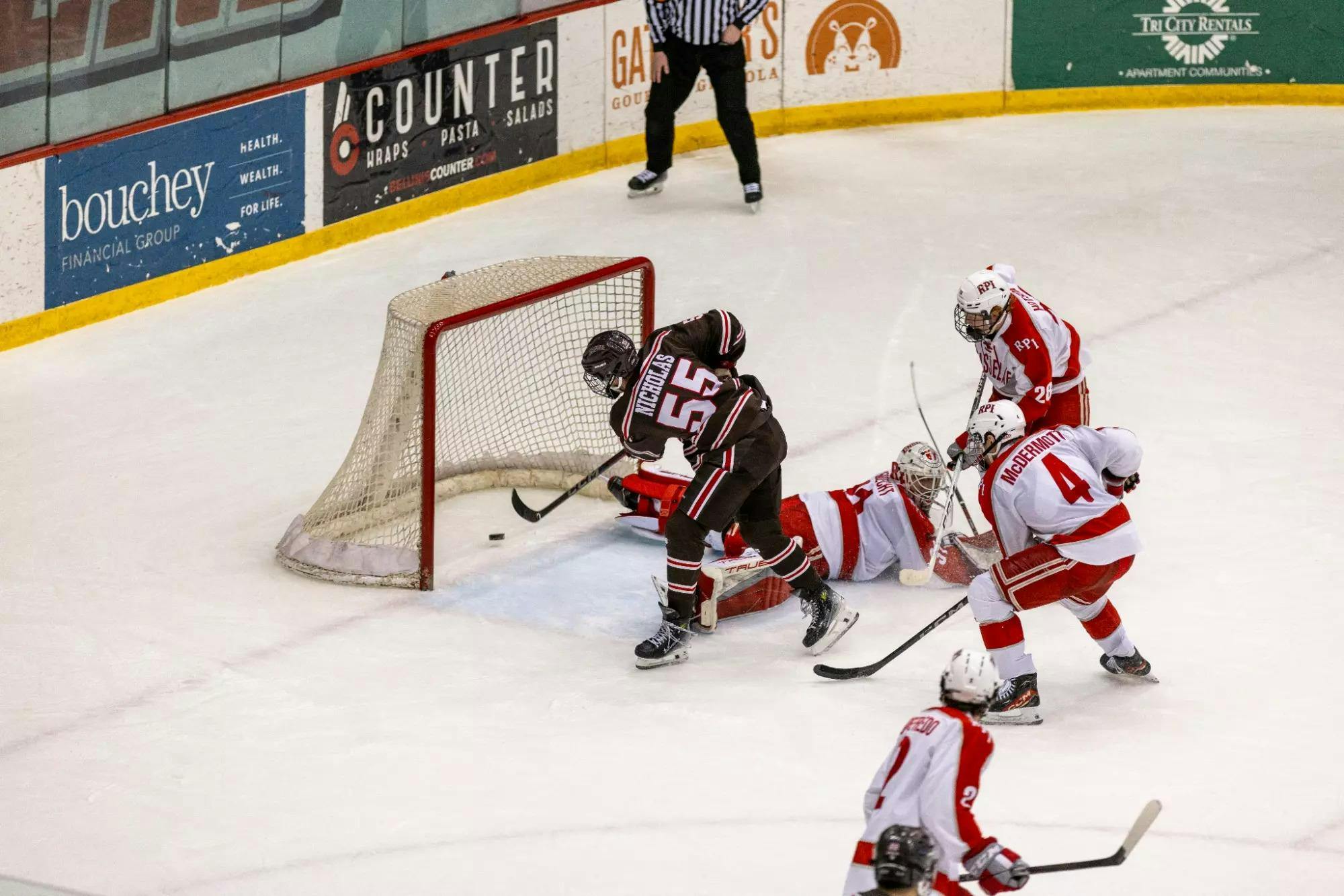 Four RPI hockey players wearing white uniforms surrounding Brown University hockey player Brian Nicholas, who is wearing a brown uniform and scoring a goal.