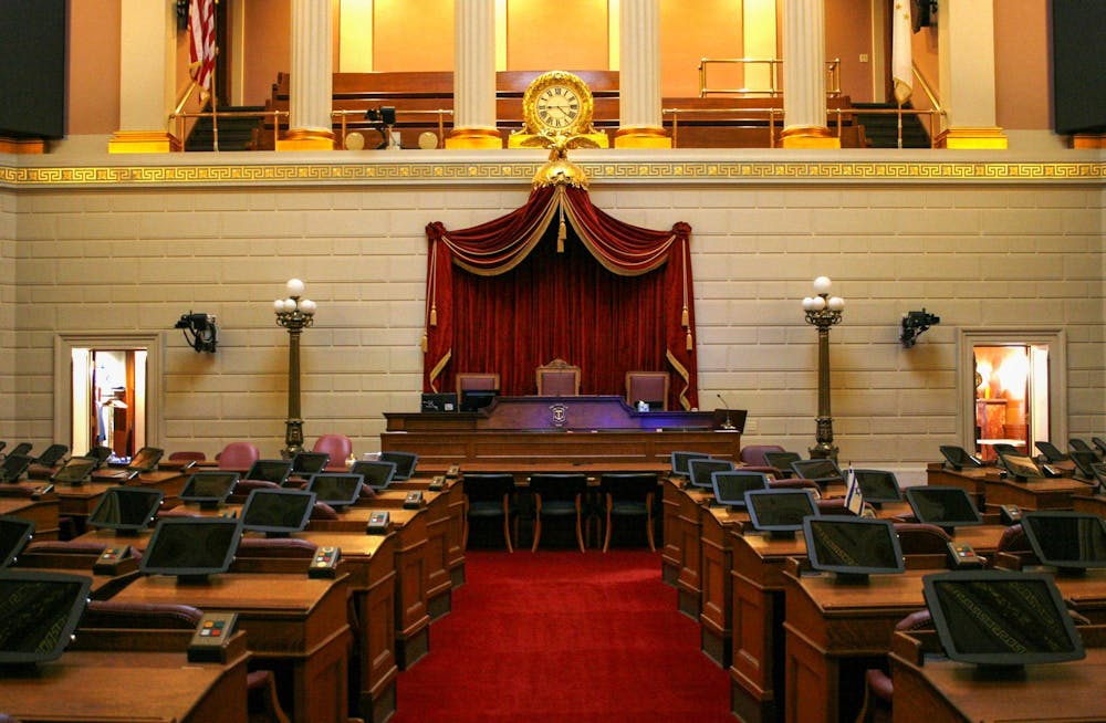 Courthouse with several wooden tables on the left and right with tablets and voting button sets. In the center is a red carpeted walkway with a large table in the center with red curtains.