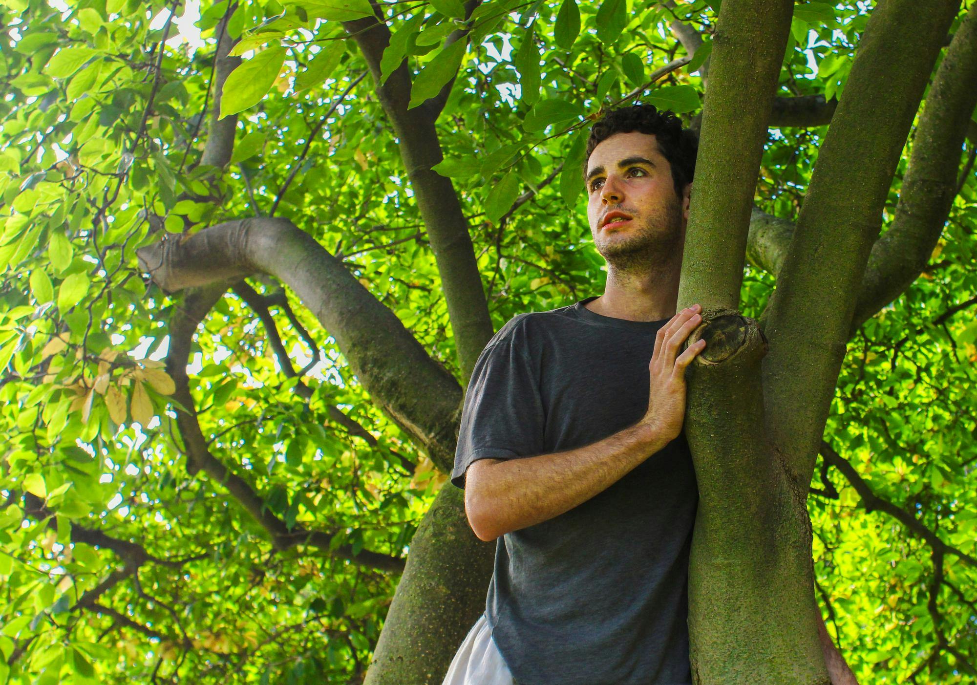  A young man with brown hair looks longingly behind a tree branch.