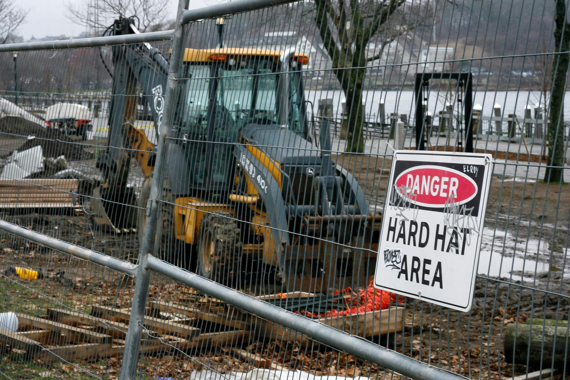 A picture of the construction occurring at the plaza at India Point Park. In the picture, there is a fence with a sign reading "Danger: Hard Hat Area," and behind the fence, there is a construction vehicle.