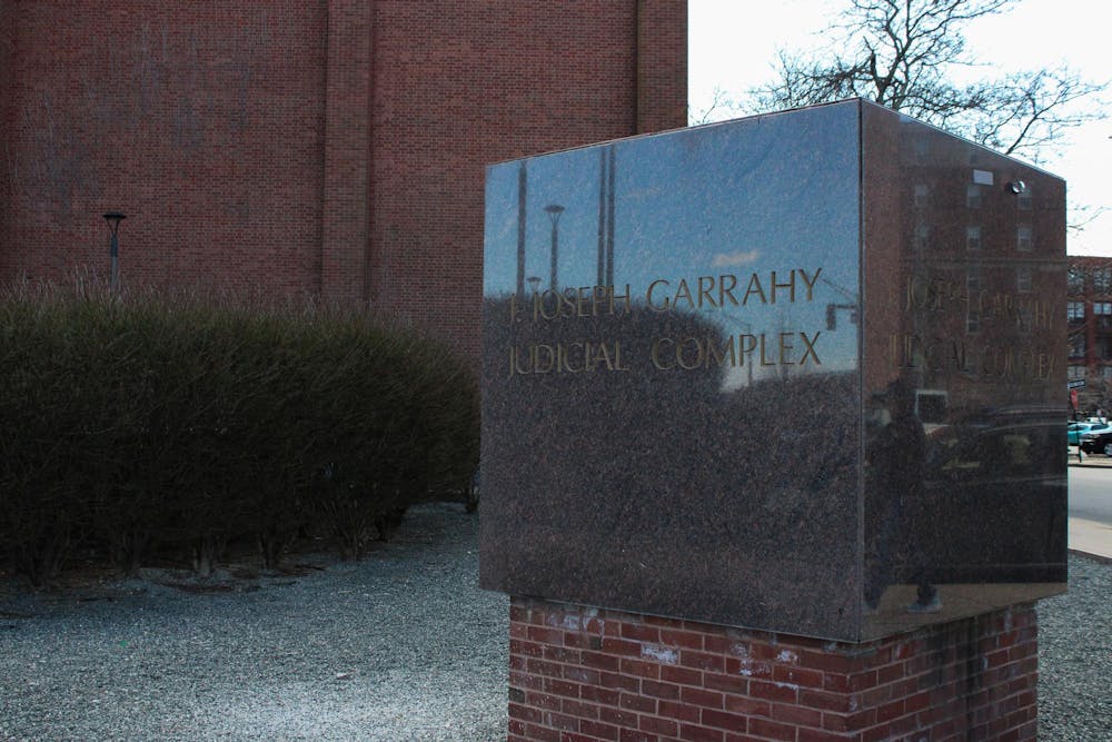 Photo of a stone cube with the words “J. Joseph Garrahy Judicial Complex” engraved in capital letters in front of a brick building and bushes. 