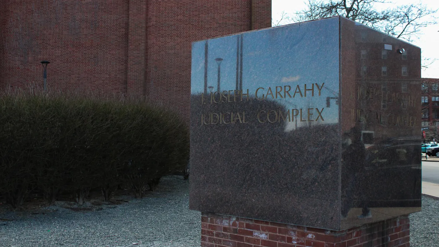 Photo of a stone cube with the words “J. Joseph Garrahy Judicial Complex” engraved in capital letters in front of a brick building and bushes.