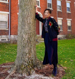 Photo of a woman leaning against a tree, her jacket embroidered with pomegranates. 