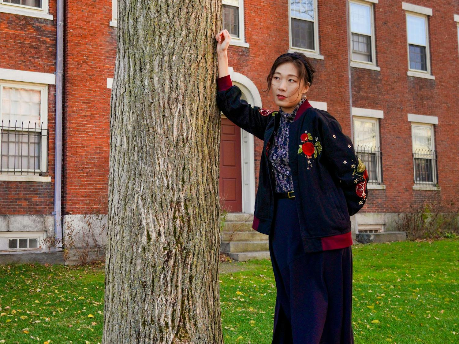 Photo of a woman leaning against a tree, her jacket embroidered with pomegranates.