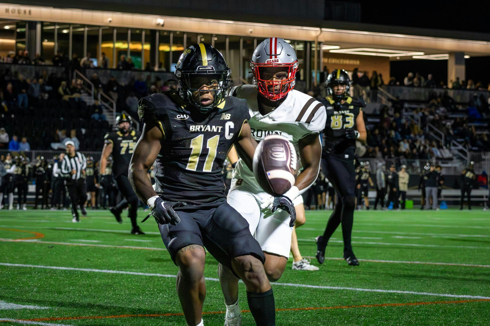 Photo of a Bryant football player and a Brown football player watching a football fall in front of them with bleachers in the background.