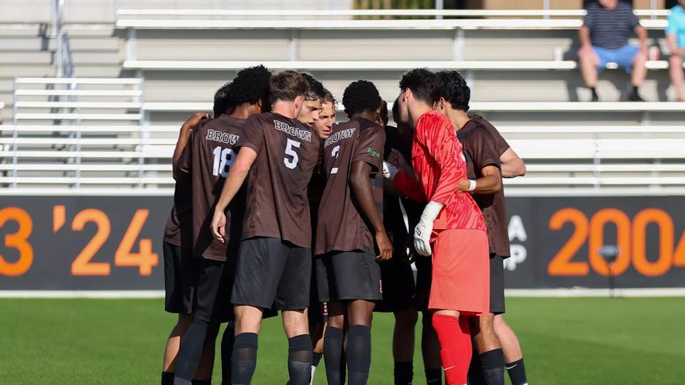 Photo of Brown men's soccer players leaning into a huddle on the soccer field.
